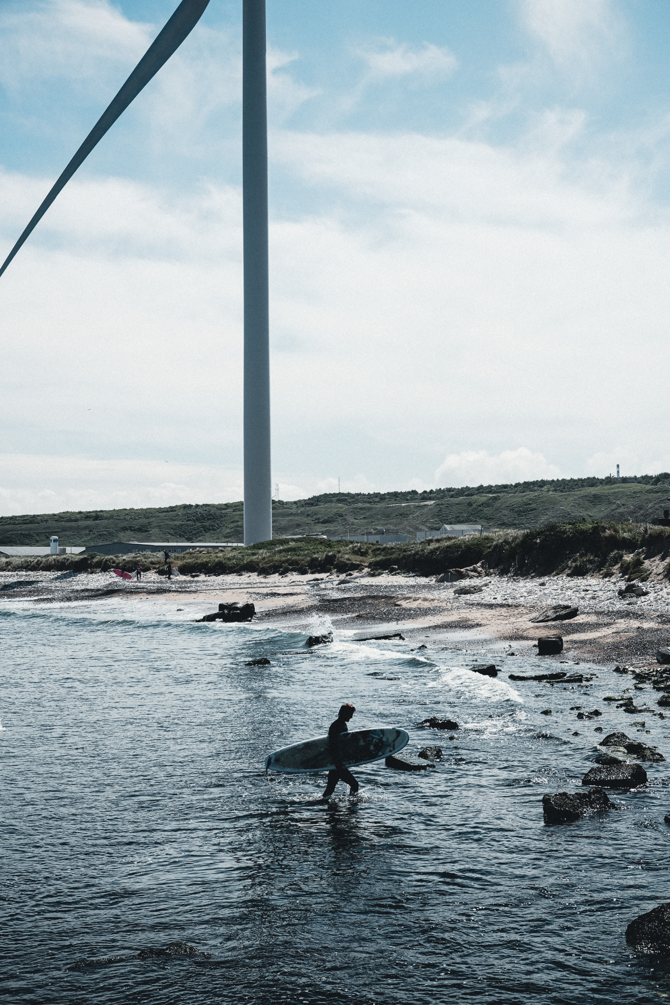Surfer geht mit Surfbrett aus dem Arm aus dem Wasser, am steinigen Strand. Im Hintergrund ist ein Windkraftwerk zu sehen Lino Albrechtsen - linoalbrechtsen.de